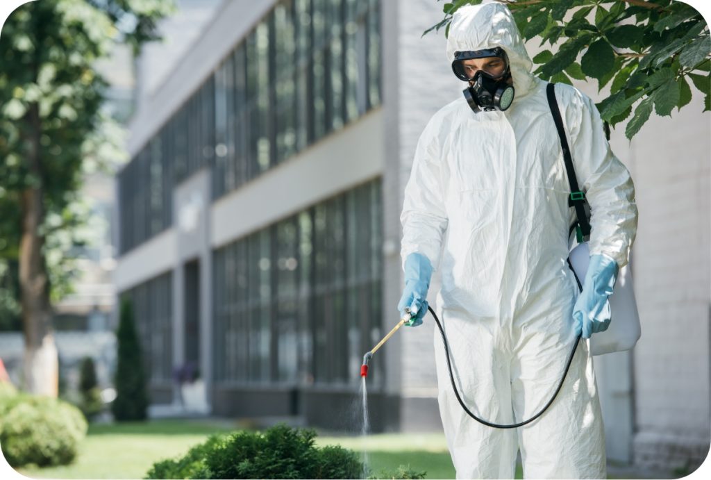 Cleaning technician performing outdoor pest control treatment with protective gear at a commercial property.